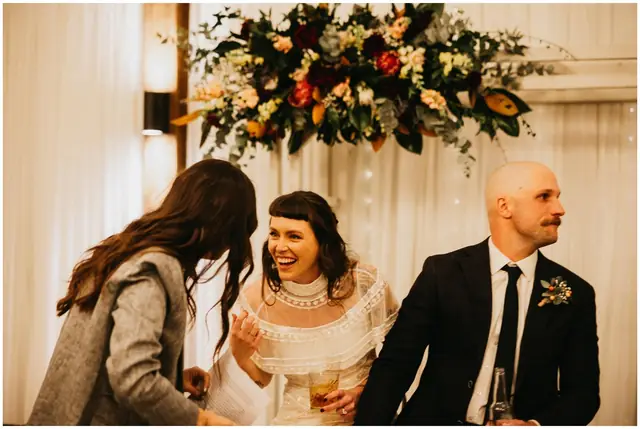 Luke & Cara at Cowbell Creek: The bride laughs with a guest beside the groom and a floral arrangement.