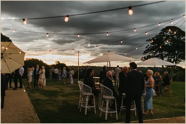 Jess & Rich at The Orchard Estate: Guests mingle outside beneath festoon lights at dusk.
